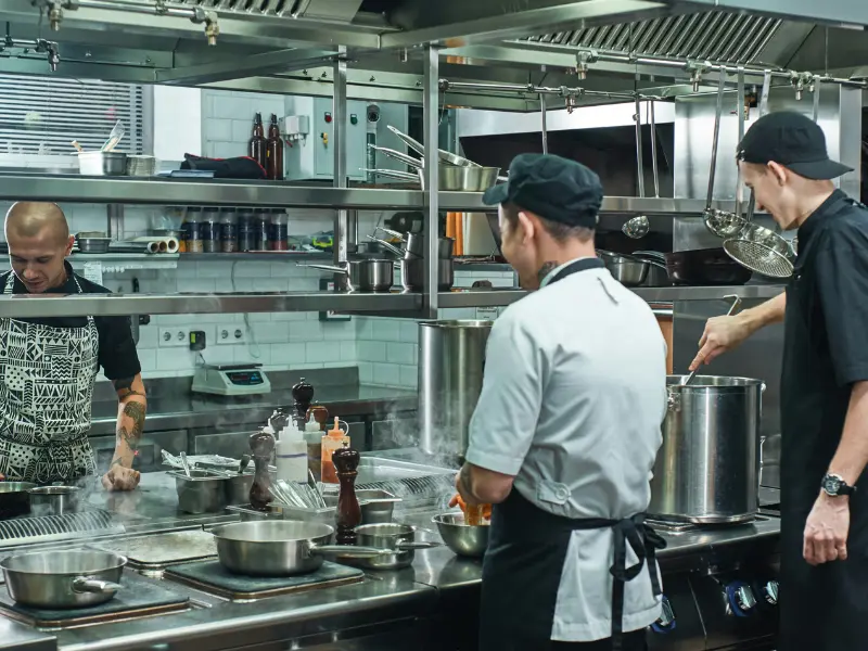 
The image shows three chefs actively working in a busy, modern commercial kitchen. The atmosphere is focused and professional, typical of a restaurant or high-volume food production setting.