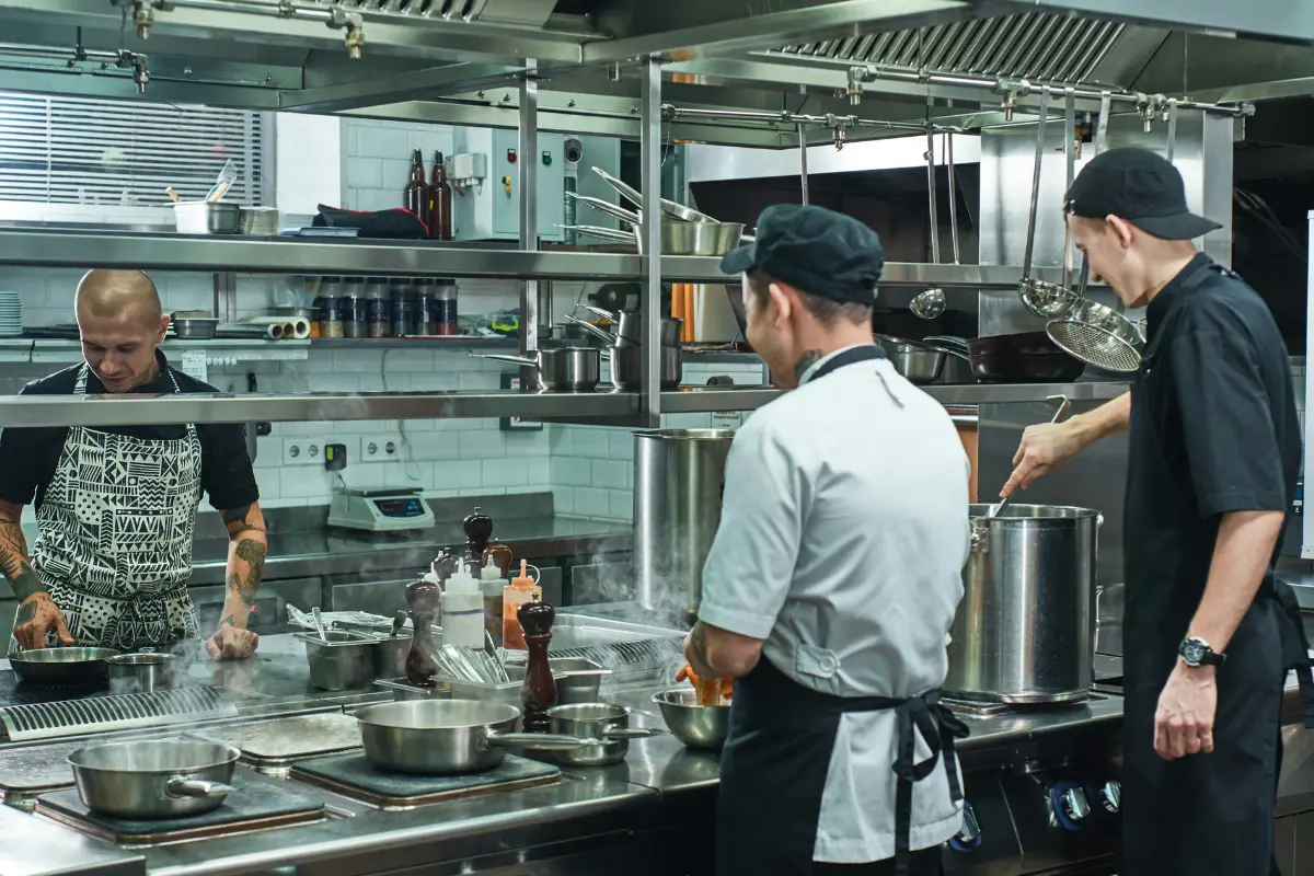 The image captures a busy moment involving three chefs working intensely on the cookline of a professional commercial kitchen.