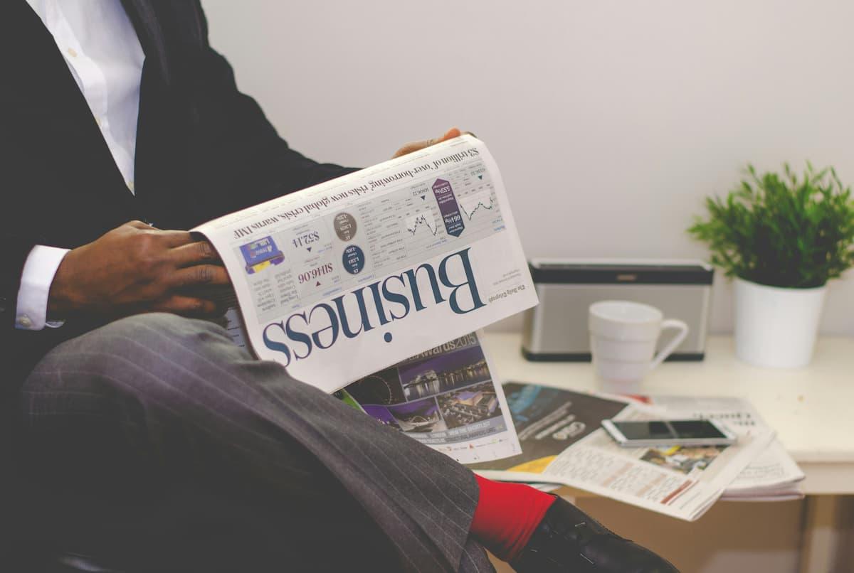 Man sitting in a suit, reading a business newspaper with a desk in the background, featuring a plant, a mug, and a smartphone.
