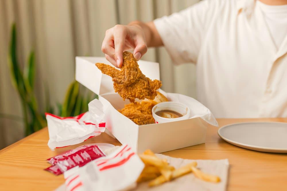 A person holding a piece of juicy fried chicken, accompanied by fries and sauce in a box