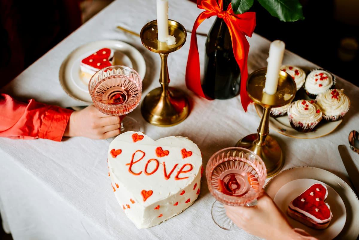 A table decorated for Valentine's Day, featuring an array of desserts like heart-shaped cookies, a cake with "love" written on it, and cupcakes topped with white frosting and red sprinkles. Two hands are visible holding glasses of pink wine, adding a romantic touch. The setting also includes brass candle holders and a bottle tied with a red ribbon.
