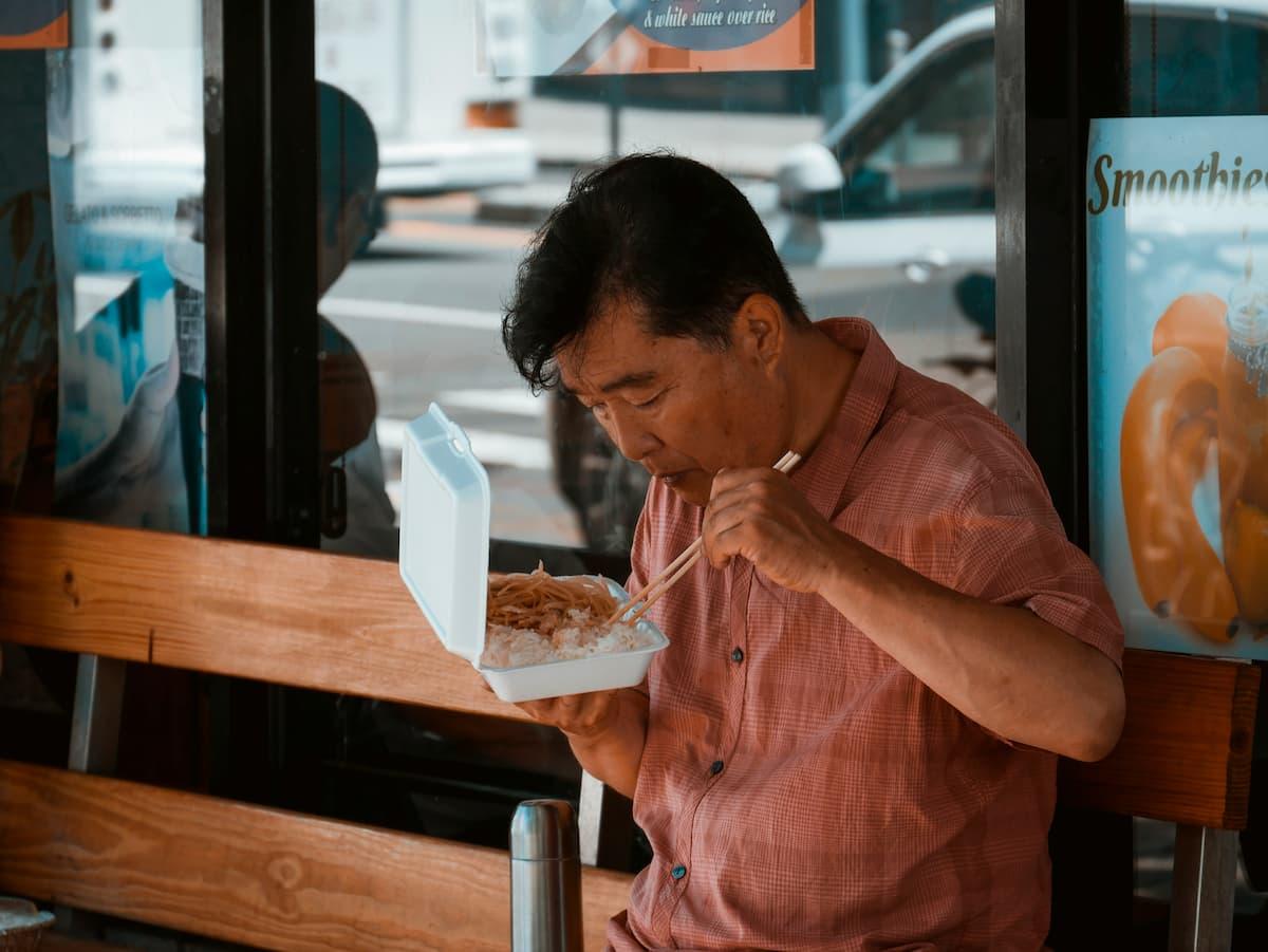 Man in a pink shirt eating from a white takeout container with chopsticks, seated on a wooden bench in an outdoor setting, with advertisements for smoothies and food in the background.