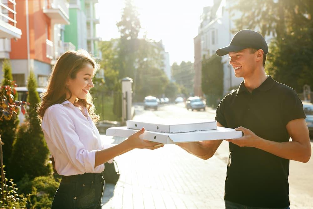 Young person receiving a pizza delivery on a sunny day. The delivery person smiles while handing over pizza boxes