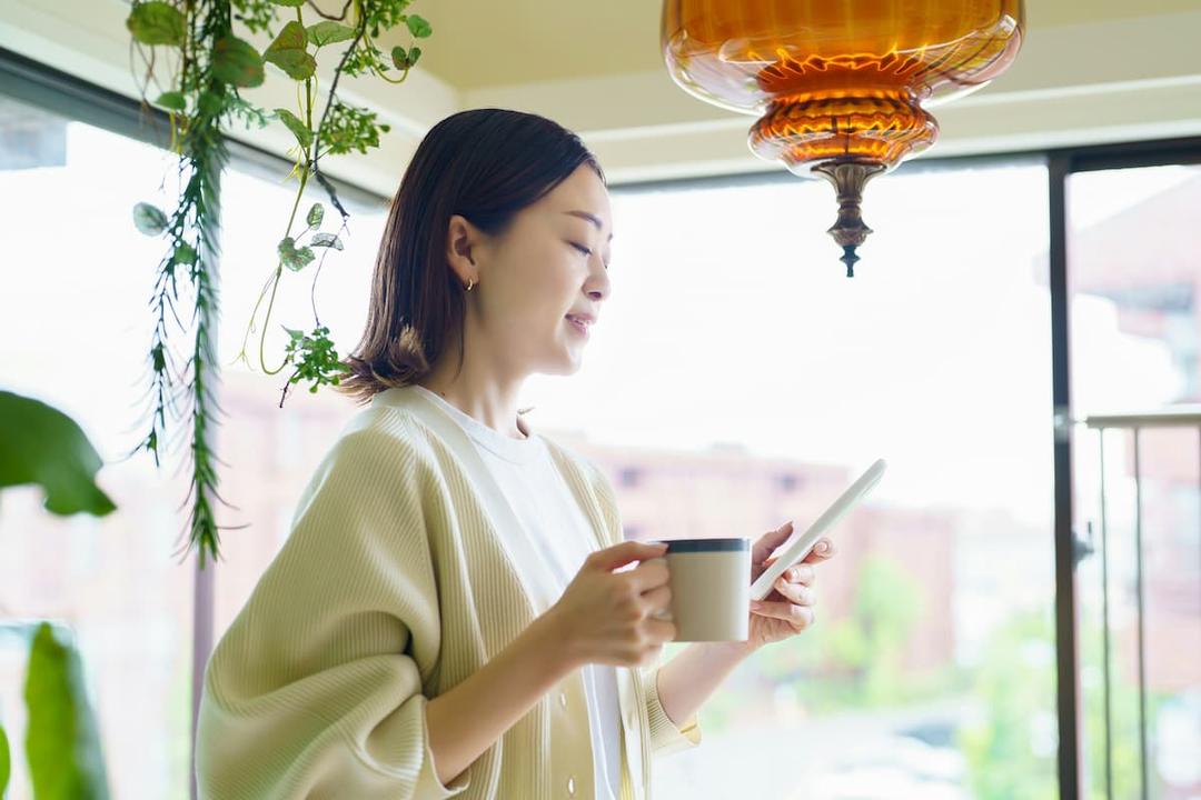 A person stands indoors near a window surrounded by hanging plants, holding a tablet while ordering food online. In the other hand, they hold a coffee mug, creating a cozy and modern atmosphere