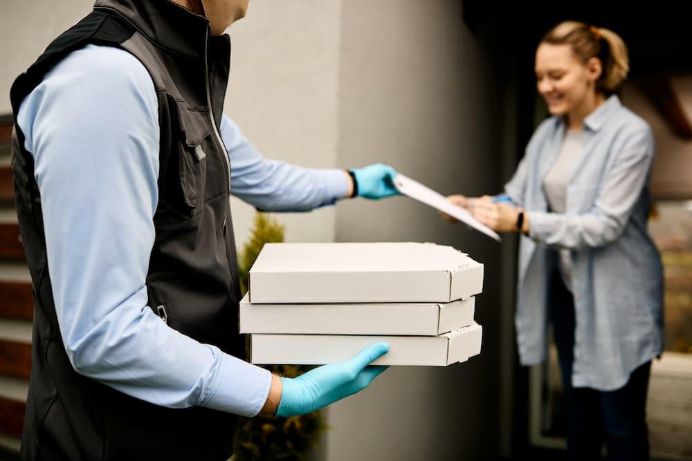 Food delivery worker wearing a black vest and blue gloves is handing over stacked pizza boxes to a customer at the doorstep.