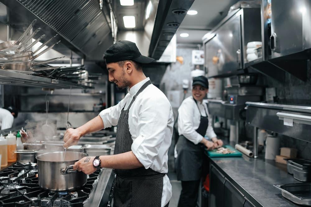 Chefs working in a professional kitchen, one chef is stirring a pot while another cook is preparing ingredients on a counter