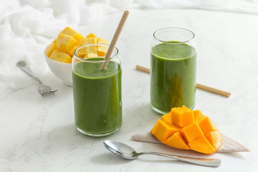 Healthy green smoothies in cups with straws, accompanied by mango cubes on a table