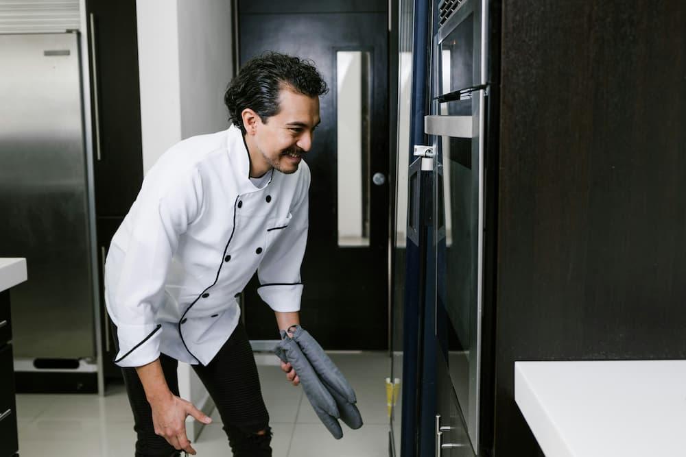 A smiling chef in a modern kitchen, wearing an apron while checking the oven
