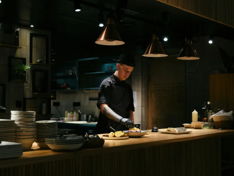 This is a photograph of a chef working at the pass or service counter of a kitchen with a modern, dark design aesthetic.