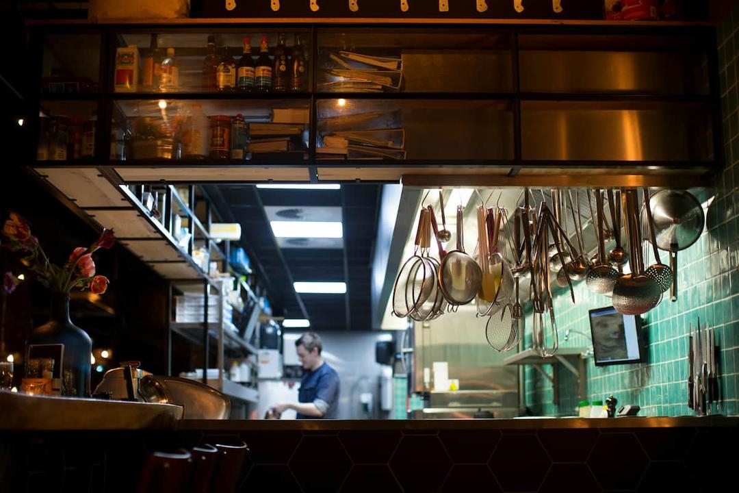 Photo of a commercial kitchen with utensils hanging from above and a chef working in the background.