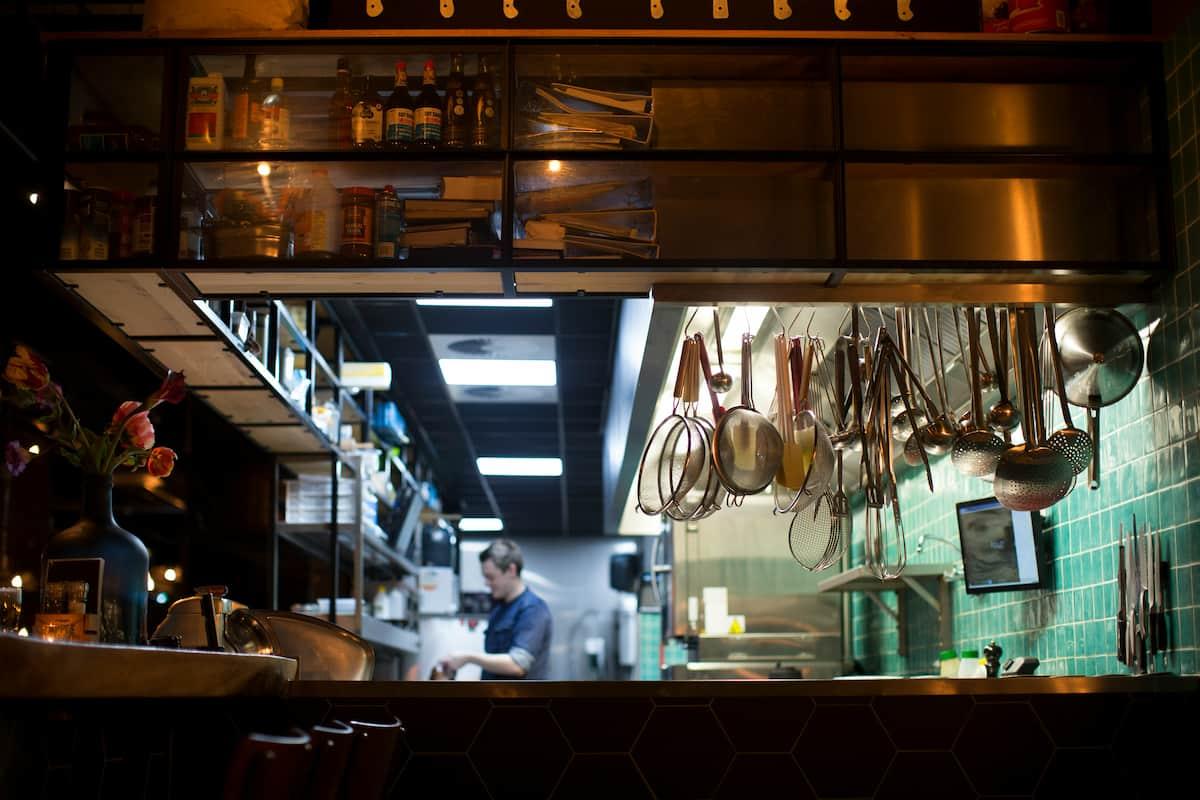 Photo of a commercial kitchen with utensils hanging from above and a chef working in the background.