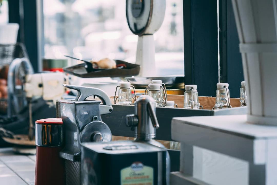 The image depicts a section of what seems to be an industrial kitchen or a restaurant's prep area. In the foreground, there are various kitchen tools, such as a metal press or grinder, condiment containers, and bottles with airtight stoppers. In the background, a food scale and a tray with what appears to be some bread or food items are visible. The overall setting suggests a functional, organized kitchen space ready for food preparation.