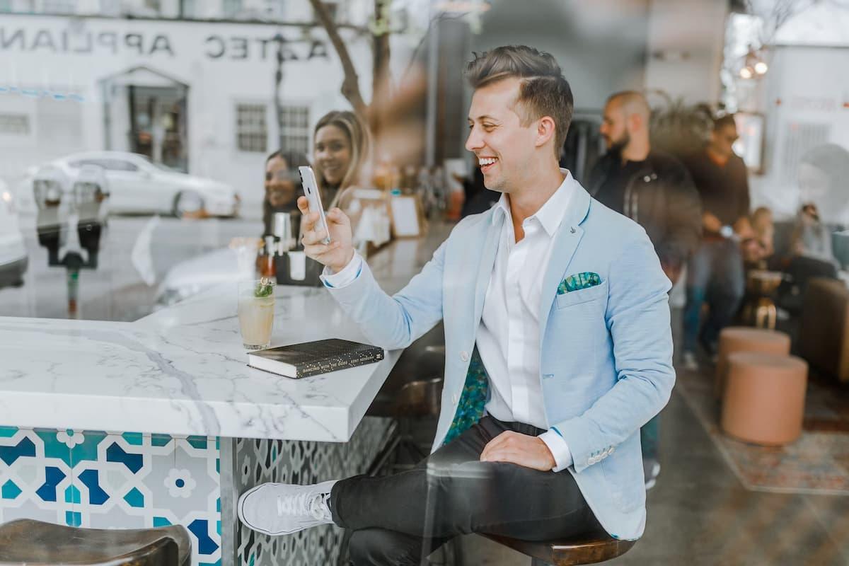 Smiling man in a light blue blazer sitting at a modern café, holding a smartphone and interacting with an app, representing customer engagement and loyalty in a restaurant setting.