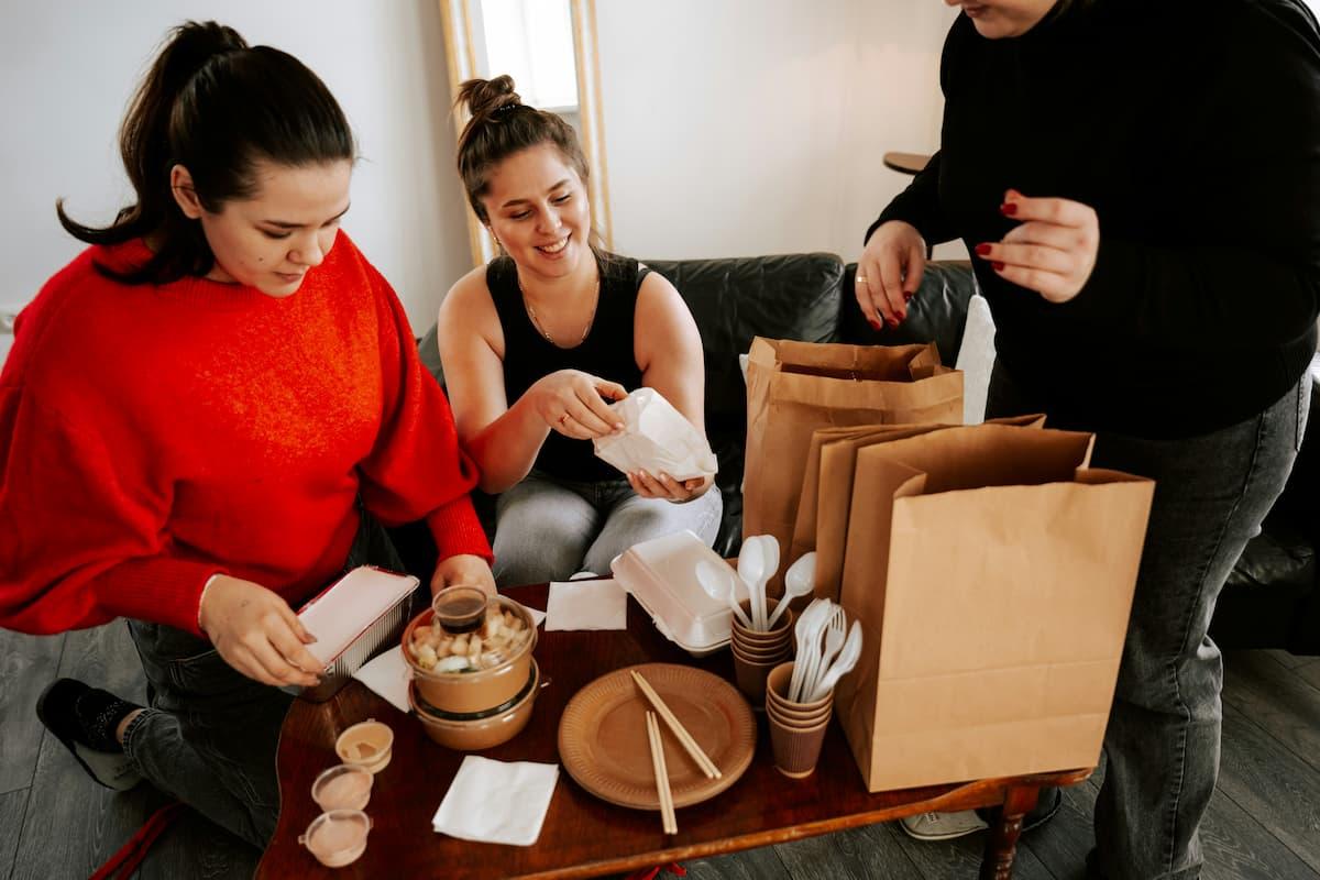 Casual gathering with pre-ordered food takeout spread across a table, featuring various takeout containers, paper bags, disposable utensils, and napkins