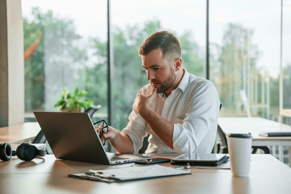 Man focused on working on a laptop in a modern office, surrounded by nature, wearing headphones, and with a mug beside him