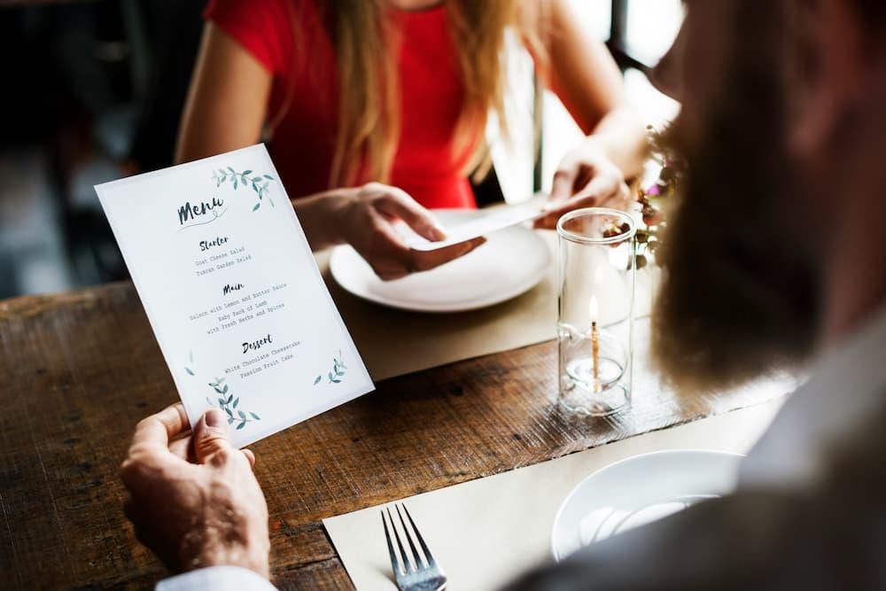 Two people seated at a restaurant table, each holding menus. The table is set with plates, cutlery, and a lit candle in a glass holder.