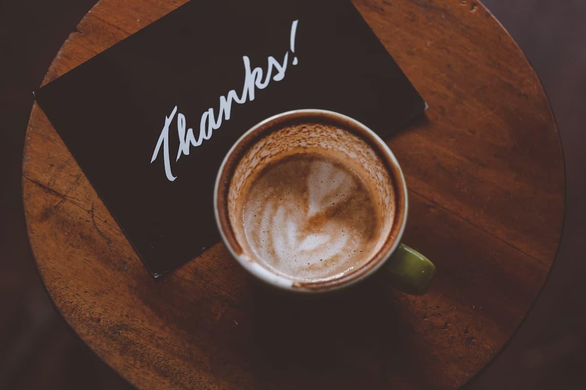 Top-down view of a half-finished latte with milk froth art in a green cup placed on a wooden table, next to a black card with the word 'Thanks!' written in white cursive.