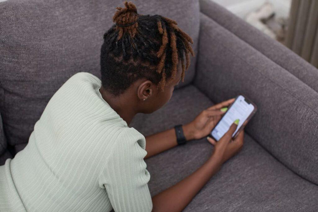 A person lying on a gray couch, using a smartphone with their hand and looking at the screen, while wearing a light green sweater.