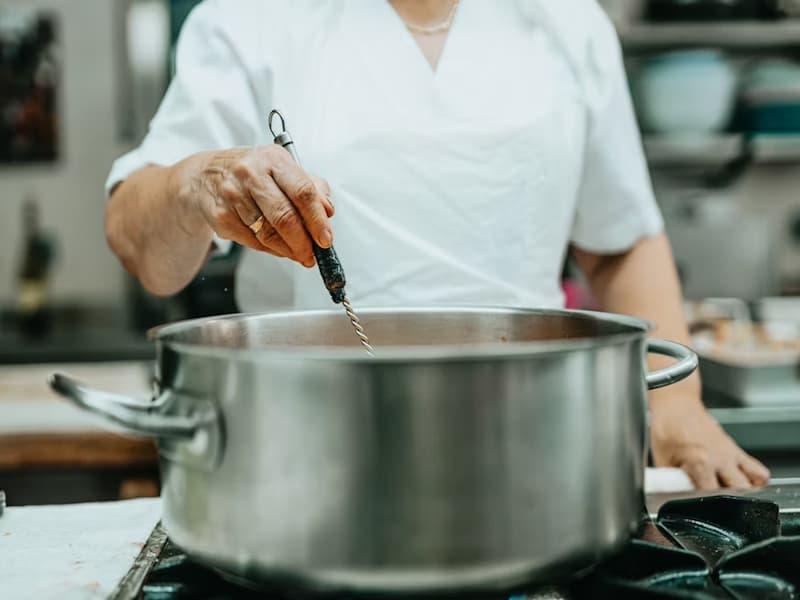 The image shows a chef in a professional kitchen stirring a large stainless steel pot on a stovetop.