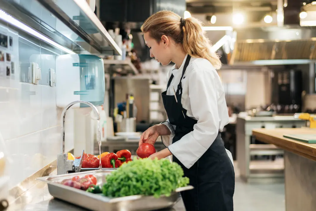 This photograph features a female chef engaged in the preparation phase of cooking in a well-lit, professional kitchen. While the image shows a high-standard commercial kitchen, its focus on efficiency and prep work is consistent with the functions of a dark kitchen or ghost kitchen hub.