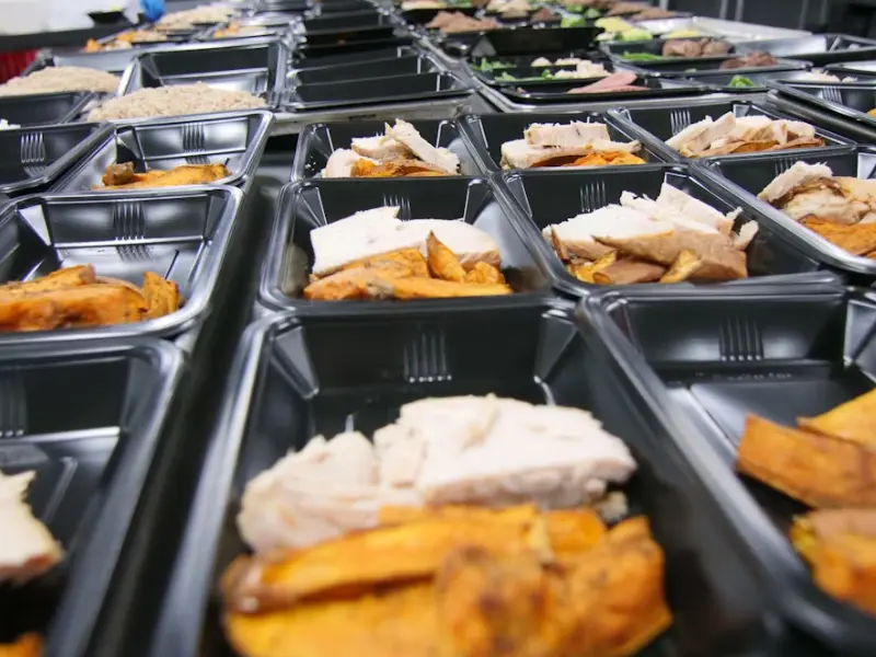 Rows of prepared meals in black containers inside a high-volume kitchen rental for large-scale food production.