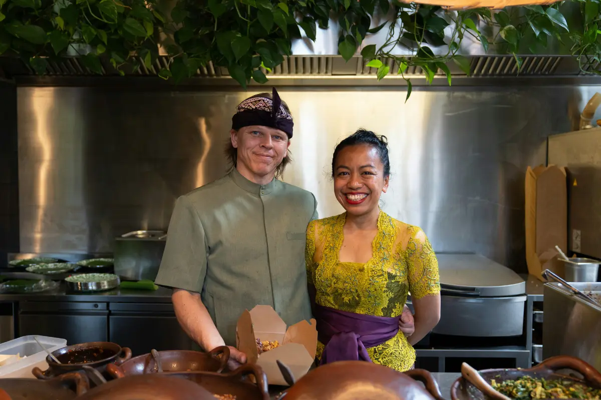 This is a portrait of a man and a woman standing in what looks like a professional kitchen. They are both smiling and appear to be a couple or business partners.