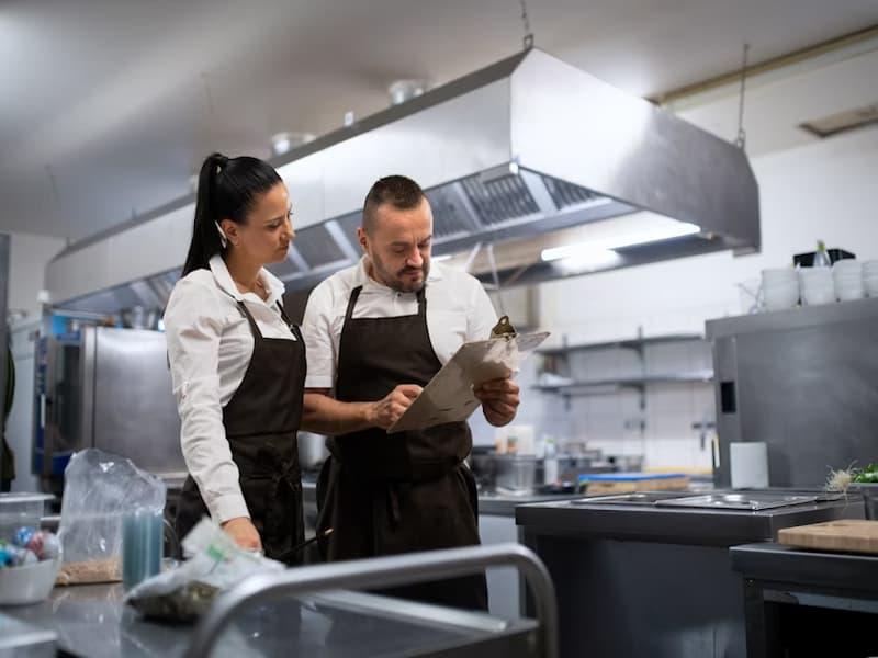 The image shows two professionals in a commercial kitchen, both wearing white shirts and dark aprons. They appear to be having a focused discussion while reviewing a clipboard, likely going over schedules, recipes, or inventory. The kitchen is clean and equipped with industrial-grade appliances, stainless steel surfaces, and ventilation hoods, suggesting a professional and organized food production or restaurant environment.