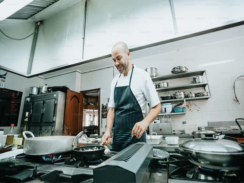 The image shows a smiling chef working in a professional kitchen. He is wearing a white chef's jacket and a dark striped apron. The chef is cooking at a gas stove, with several pots and pans in use.
