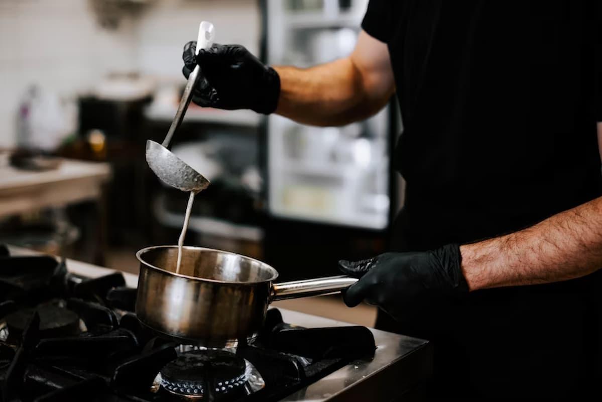 The image shows a person cooking in a professional kitchen. They are holding a stainless steel pot over a gas stove with one hand, while using a ladle to stir or pour a liquid mixture, possibly soup, sauce, or batter. The individual is wearing black gloves, which suggests a focus on hygiene and safety.