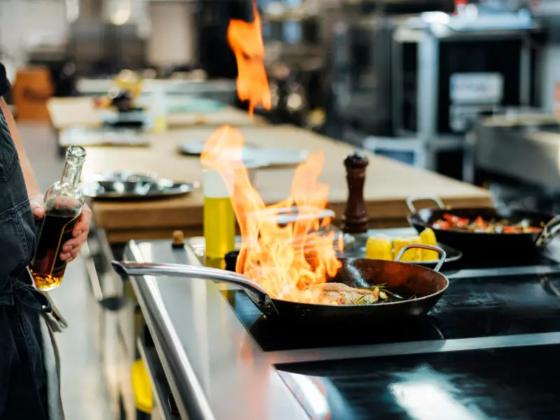The image captures a close-up, dynamic moment of flambé cooking in a professional kitchen environment.
