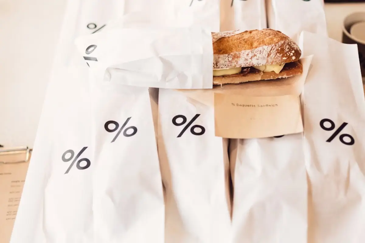 The image shows several white paper bags neatly arranged, each printed with a black "%" symbol. One of the bags is partially open, revealing a sandwich made with a crusty baguette, sliced cheese, and possibly some meat or filling.