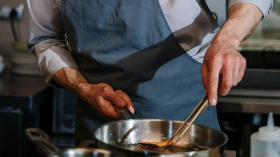 A close-up of a chef cooking in a professional kitchen. The chef, wearing a gray apron and long-sleeve shirt, uses metal tongs to handle food in a stainless steel frying pan on the stove.
