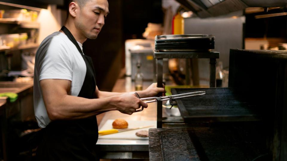 He is using a pair of long metal tongs or a specialized grill tool to adjust a heavy, blackened grill grate or tray inside a large industrial oven or broiler.