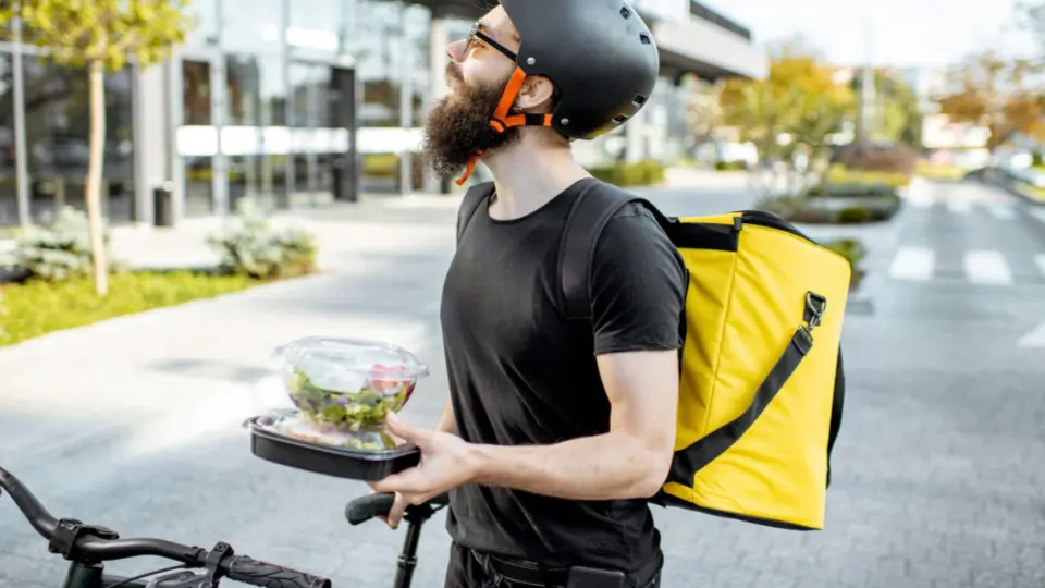 This image features a delivery courier standing outdoors on a sidewalk next to his bicycle.