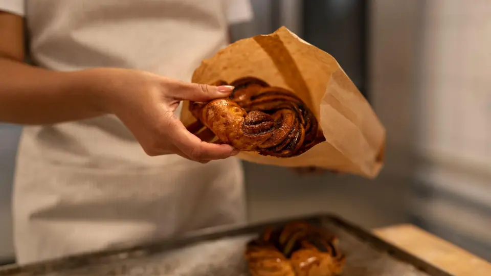 This photo shows a close-up, action shot of a bakery worker placing a fresh pastry into a brown paper bag.