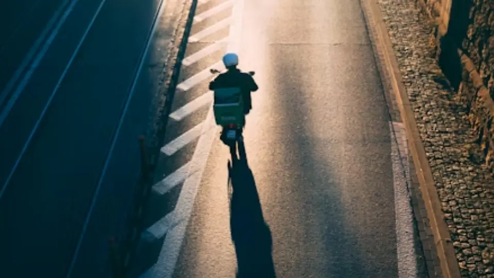 The image presents an aerial view of a delivery person on a scooter, captured from behind as they travel down a road.