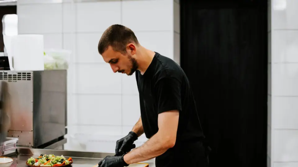 This image is a portrait of a male chef working in a kitchen. He is focused on his task, with a thoughtful expression as he cuts ingredients with a knife.