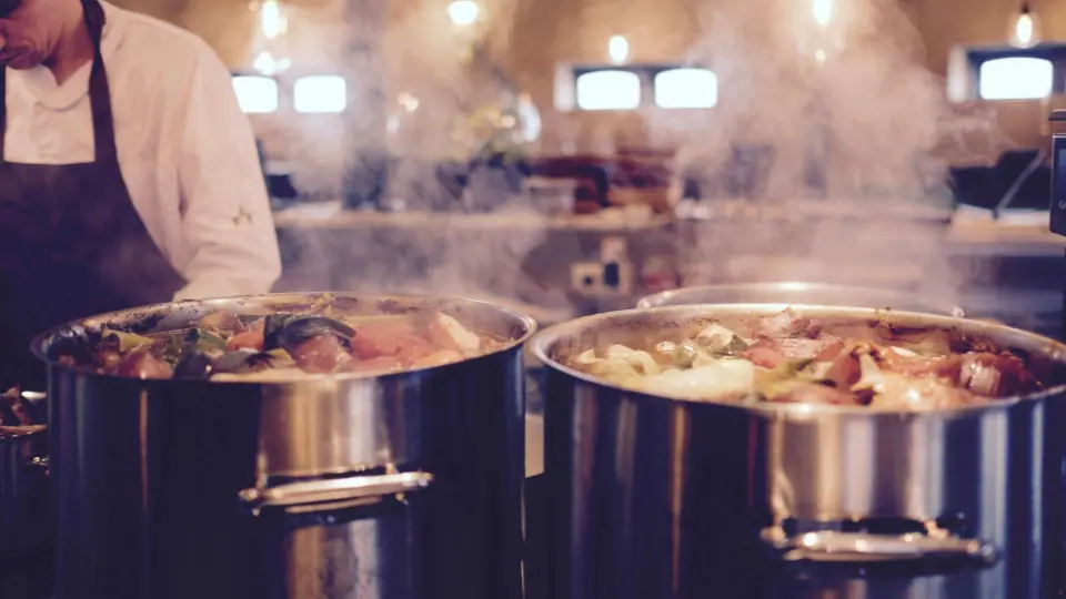 Two large industrial cooking pots filled with food are shown steaming heavily in a professional kitchen.