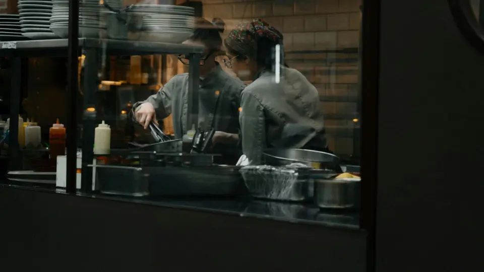 A professional kitchen is seen through a glass window, where two chefs are working side by side.