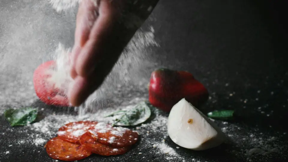 A hand sprinkling flour over ingredients on a dark surface. Slices of pepperoni, fresh basil leaves, a red tomato, and a piece of mozzarella cheese are arranged on the table.