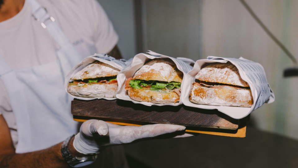 A food service worker wearing gloves and a white shirt holds a wooden serving board with three freshly made sandwiches.