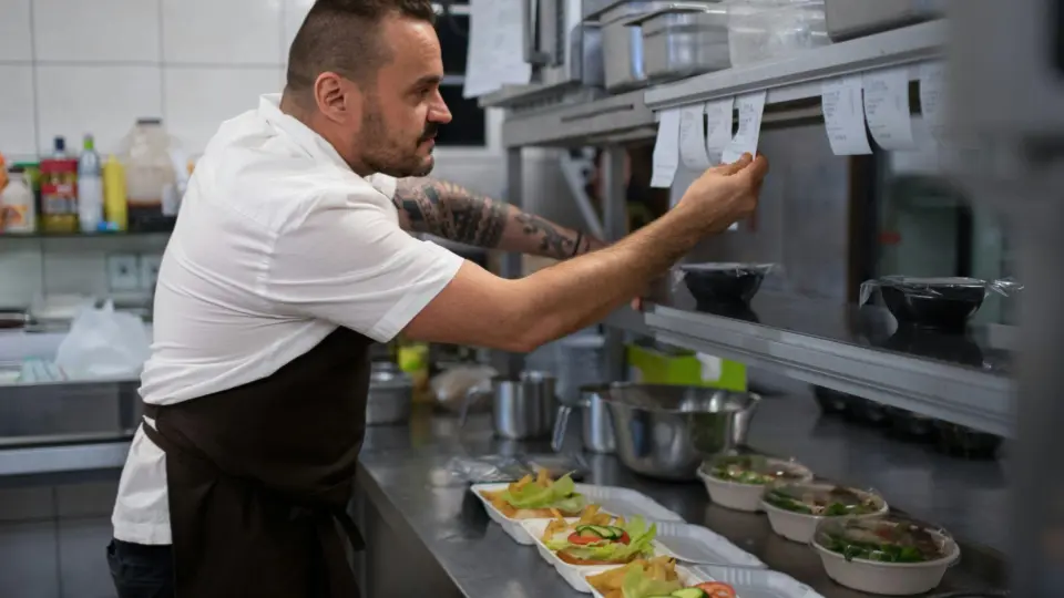 A cook wearing a white shirt and dark apron stands inside a commercial kitchen while organizing printed order tickets attached to a metal rail.