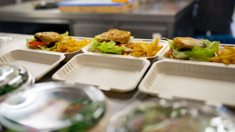 The image shows several open takeaway food containers arranged on a stainless-steel counter in a professional kitchen.