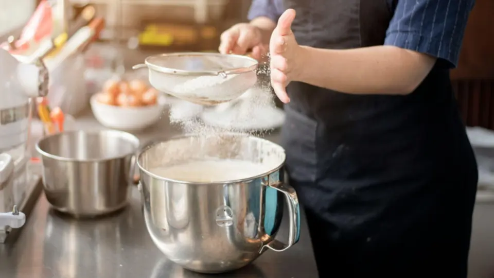 This image captures an action shot of a baker preparing a recipe, likely for bread or a delicate cake, in a professional or high-end home kitchen.