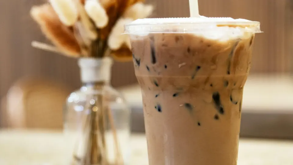 This image is a close-up of a refreshing-looking iced latte or cold brew coffee served in a clear, disposable plastic cup with a lid and a straw.