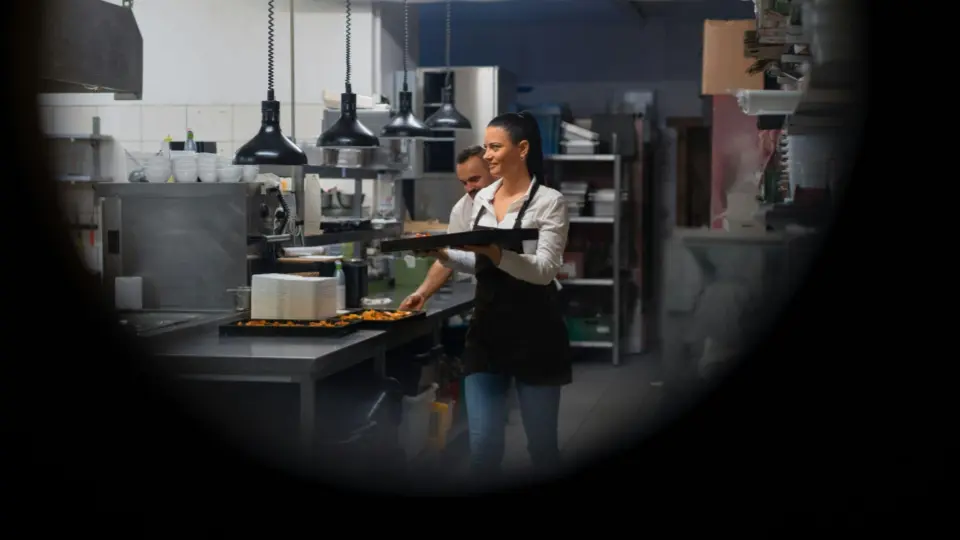 The image shows a restaurant kitchen during service, with a female server or chef wearing a white shirt and black apron carrying a tray of plated food.