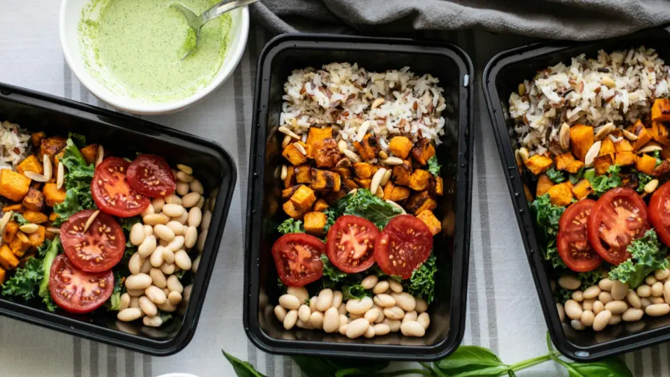 This is a top-down (flat lay) photograph showing three identical, black plastic meal prep containers. They are arranged neatly on a striped linen tablecloth.