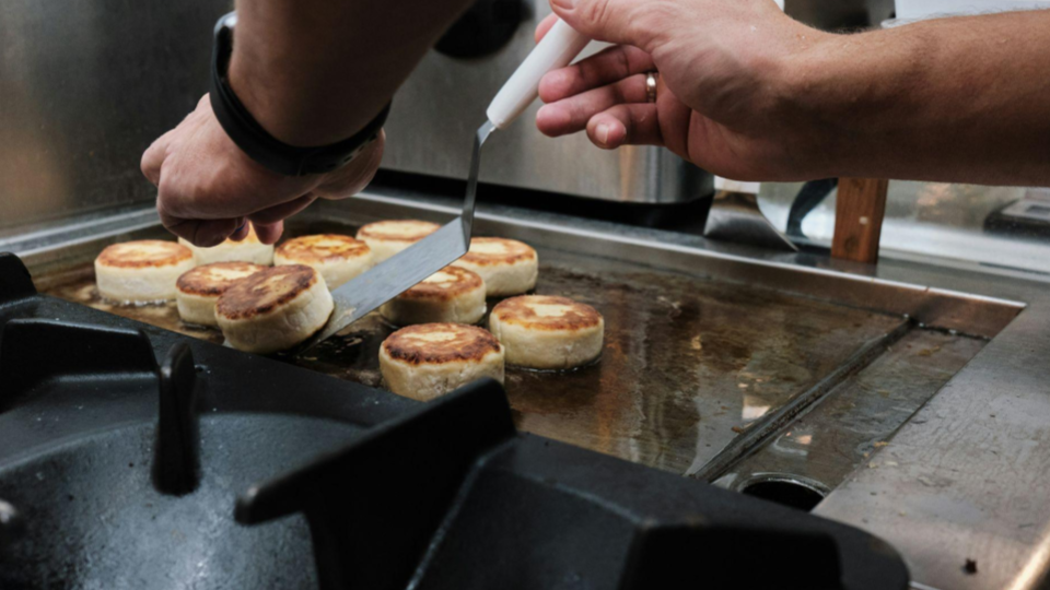 This image captures a close-up, action-oriented moment of a chef using a flat metal spatula to flip several small, circular items on a commercial griddle.