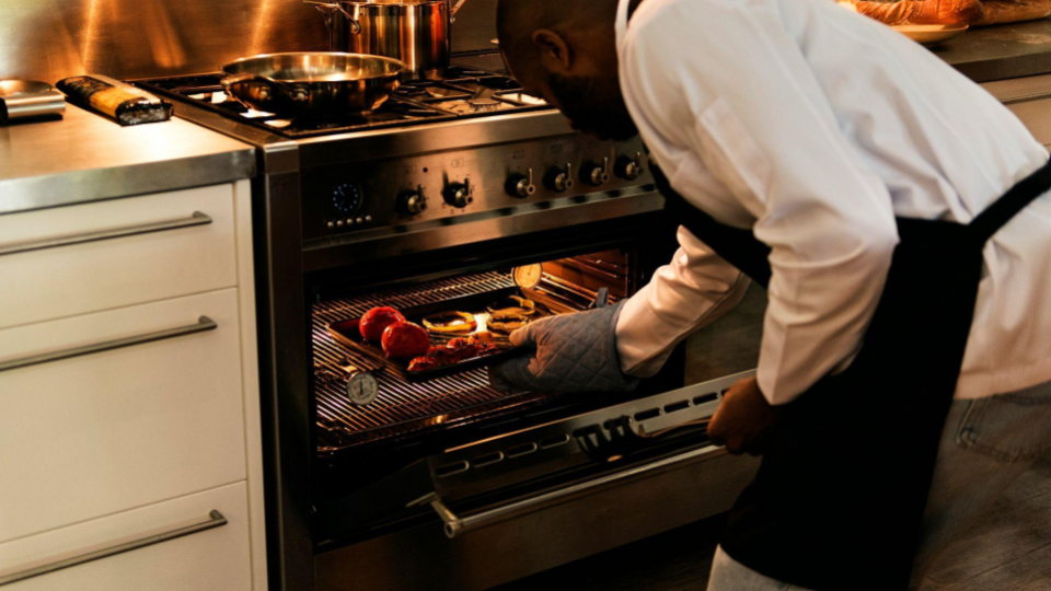 A man dressed in a professional white chef’s coat and a black apron is shown in a dynamic pose.