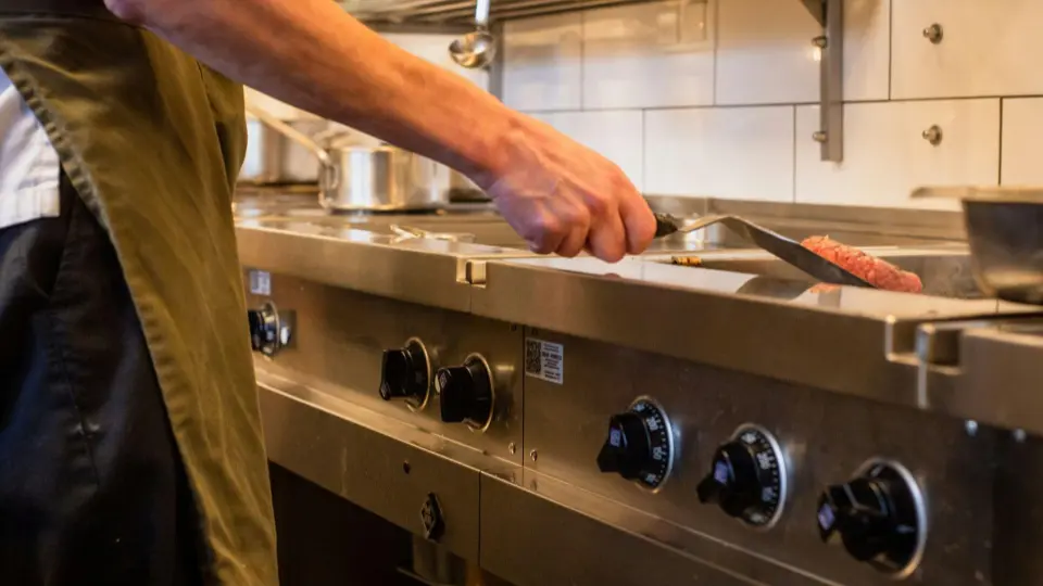 A chef is using a slotted metal offset spatula to carefully flip or move a thick, seasoned beef burger patty on a high-heat industrial griddle.